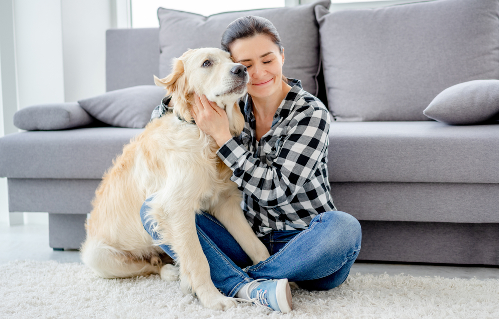 A woman sits on the floor in front of a gray couch, lovingly hugging a Golden Retriever, capturing a peaceful moment that may follow care from Emergency Vet Fleming Island