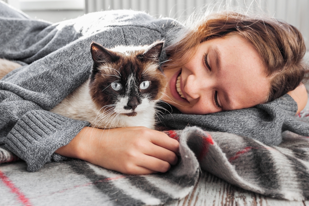 Smiling woman cuddling her cat after treatment at Emergency Vet Palatka FL A happy woman in a gray sweater lies on a blanket next to her cat with striking blue eyes and a dark face, showing warmth and relief after care from Emergency Vet Palatka FL