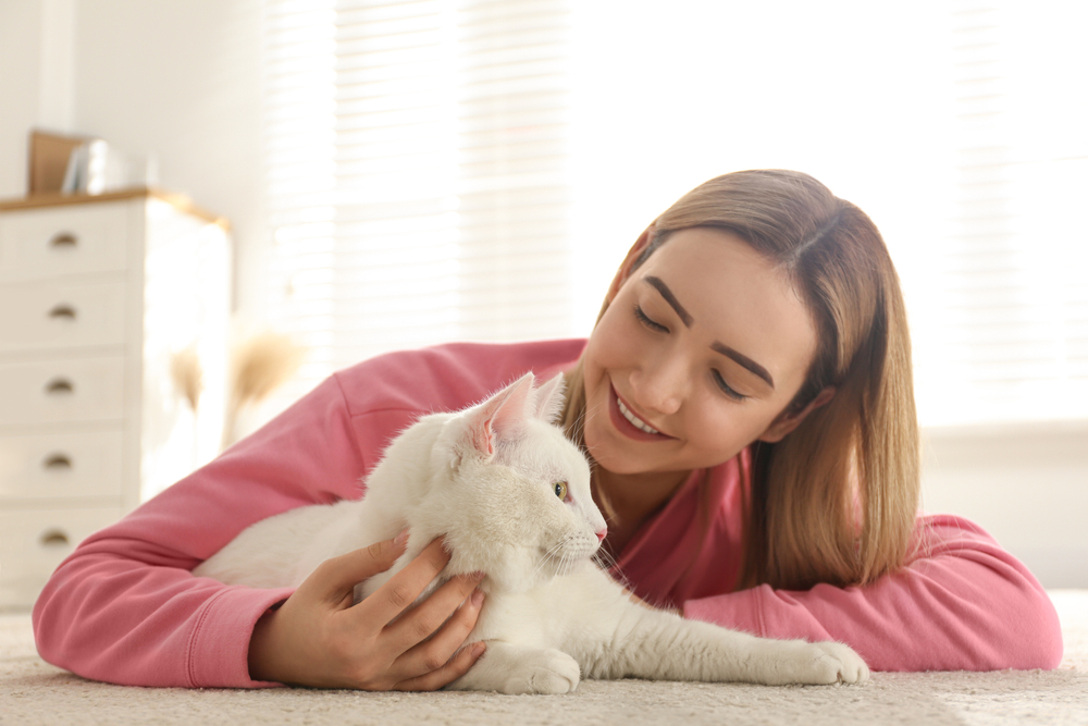 Woman smiling at her white cat after visiting Emergency Vet Palatka FL A young woman in a pink sweater lies on the floor, gently petting and smiling at her white cat, capturing a comforting moment after care from Emergency Vet Palatka FL