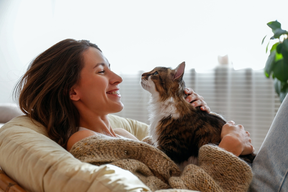 Woman Relaxing with Her Cat - EcoVets Specialists & ER A woman in a cozy sweater reclines in a chair, smiling as she holds and gazes at her fluffy cat. - EcoVets Specialists & ER